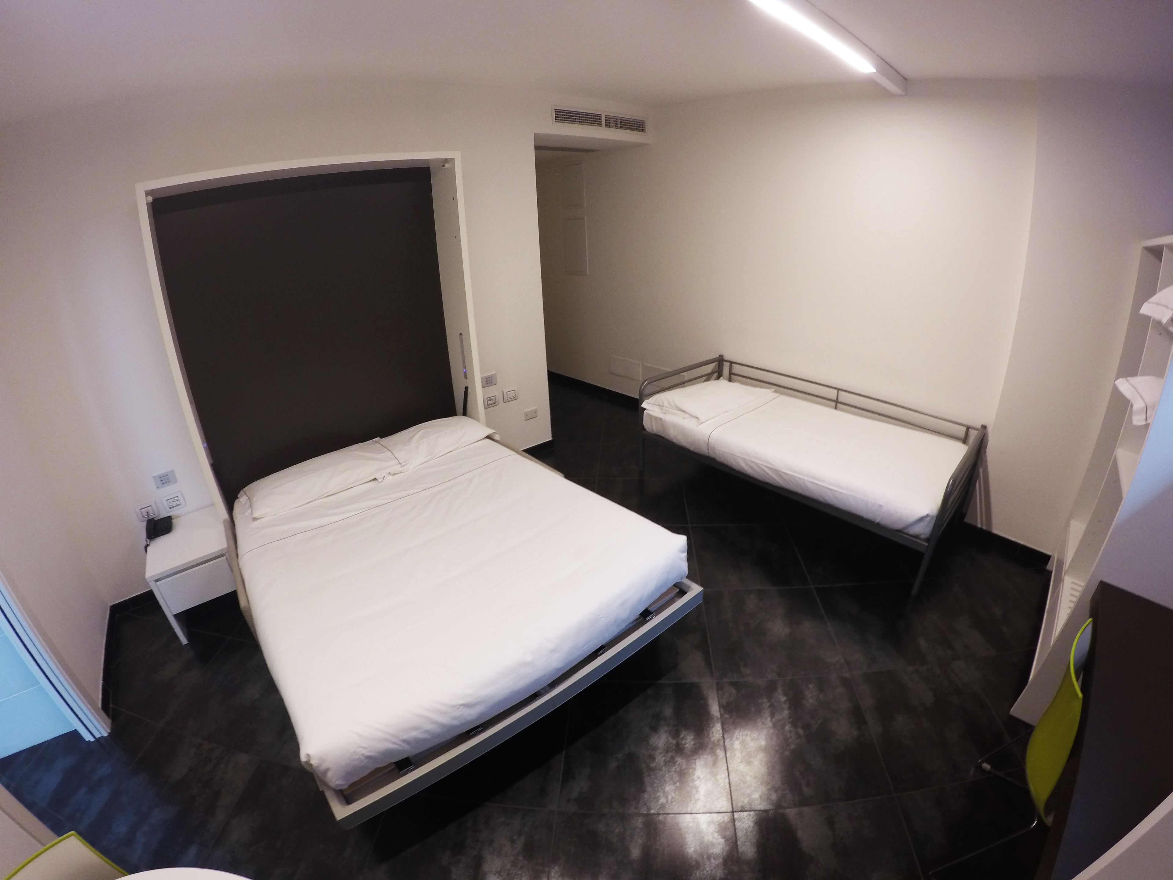 Hotelkamer met een groot bed, een kleiner bed, een nachtkastje, een stopcontact en een luchtbeheersingsunit aan de muur.