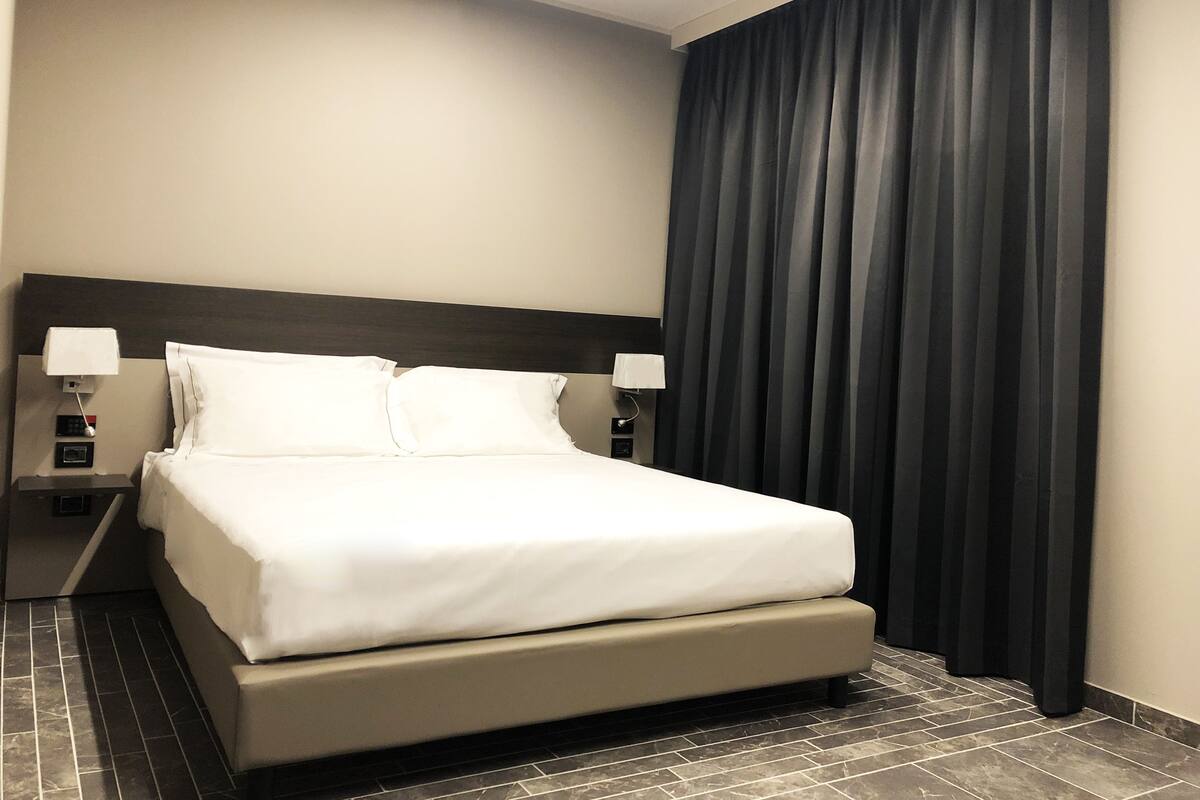 Une chambre d’hôtel avec un grand lit, deux tables de chevet avec des lampes, des rideaux sombres et un sol carrelé.