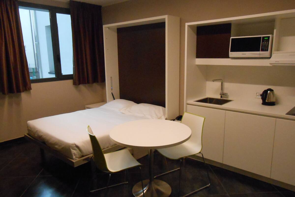 Une chambre d’hôtel compacte comprenant un lit, une petite table ronde avec deux chaises, un four à micro-ondes, une bouilloire et un évier.