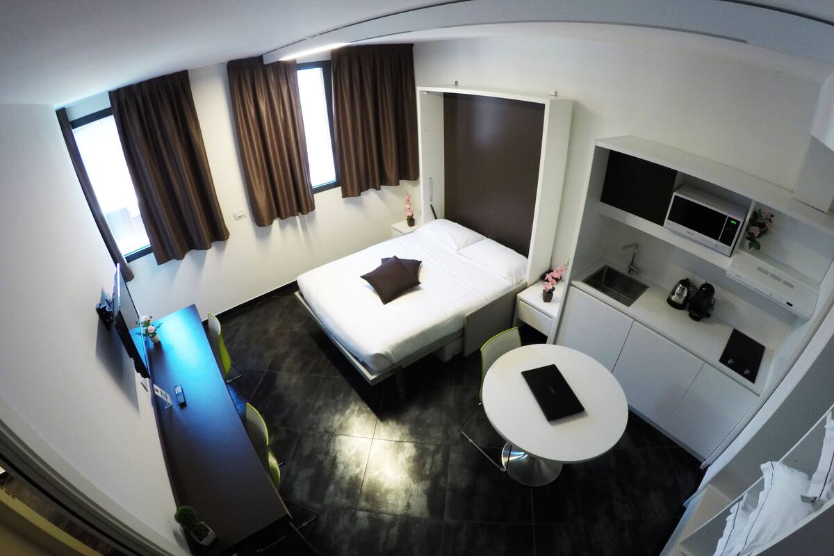 Une chambre d’hôtel de taille réduite, comprenant un lit, un petit bureau, une table ronde, un coin cuisine et une fenêtre avec des rideaux.