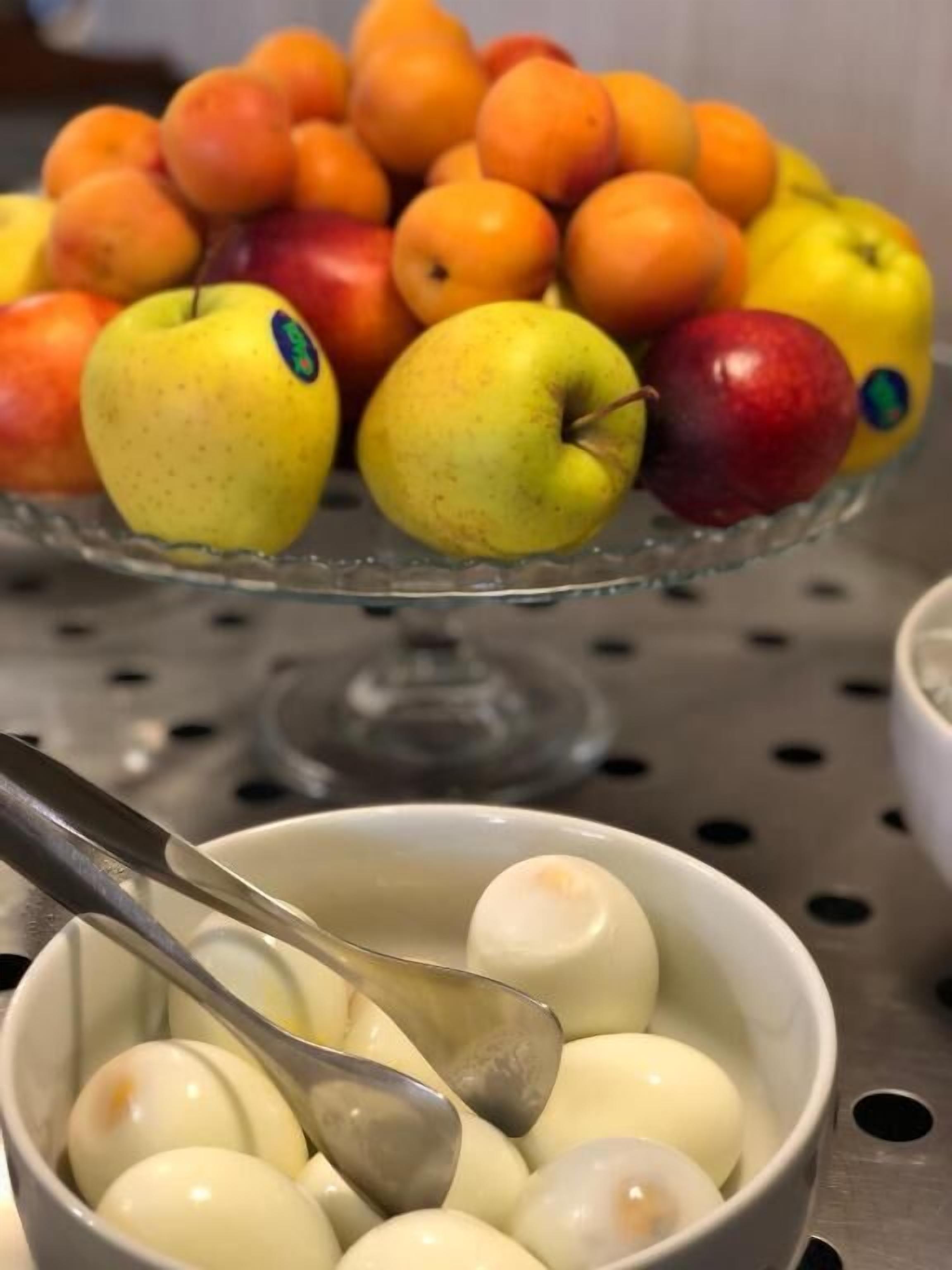 Un bol d’œufs durs, accompagné d’une cuillère, et une présentation de fruits avec des pommes et des oranges en arrière-plan.