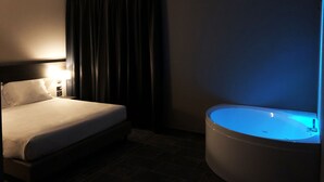 Habitación de hotel con cama, mesita de noche con lámpara, cortina oscura y bañera con iluminación azul.