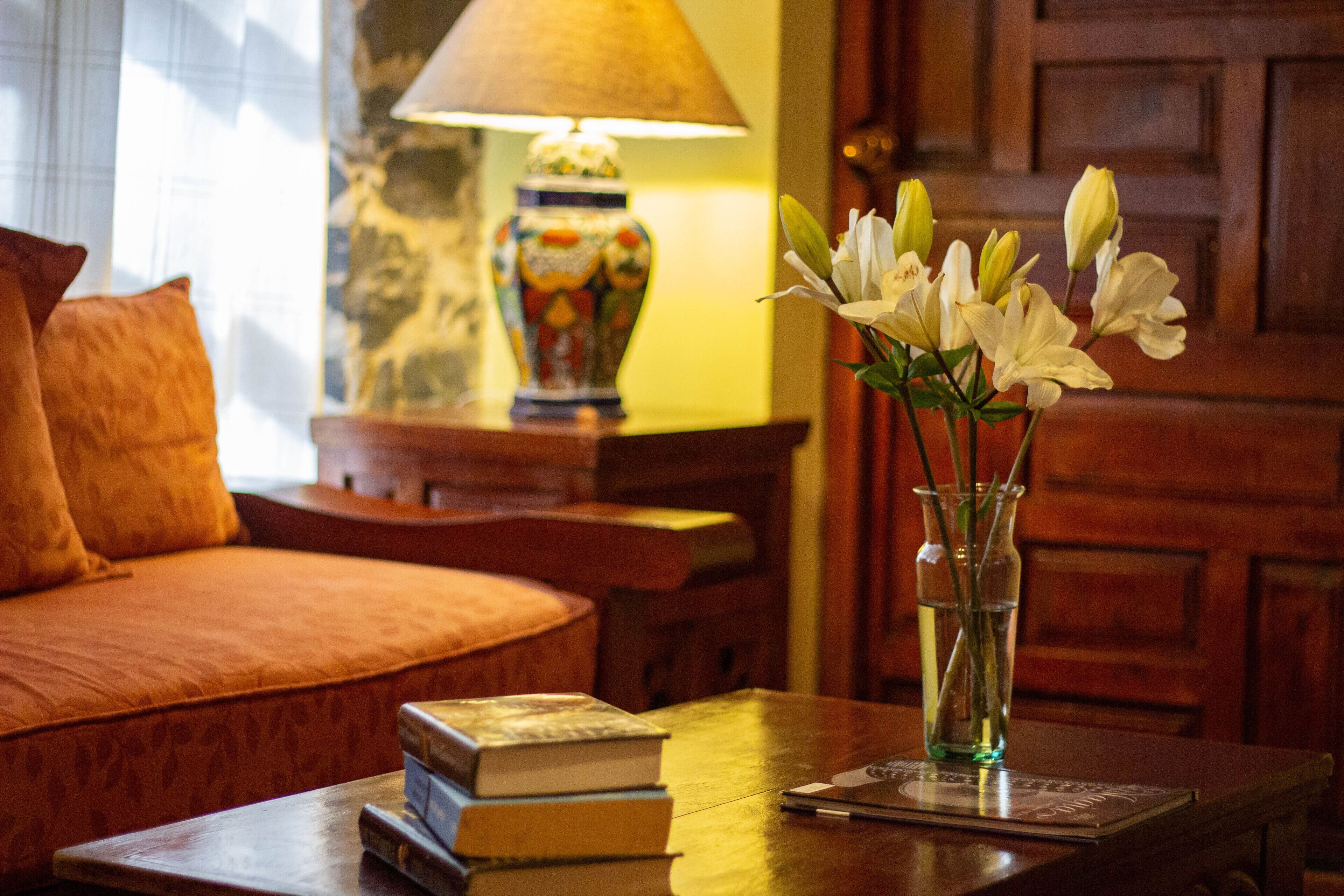 Una sala de estar acogedora con un sofá, una mesa con libros y un jarrón de flores, y una lámpara al fondo.