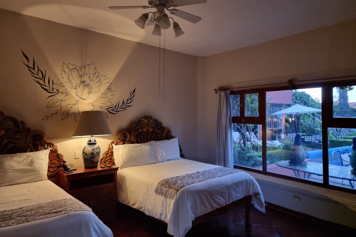 Quarto de hotel com duas camas, um ventilador de teto, um mural decorativo, um abajur e uma janela com vista para o exterior.
