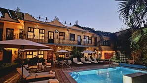 A pool area with lounge chairs and umbrellas, a clock on the building, and a staircase leading to the upper level. - Hacienda de las Flores (San Miguel de Allende)