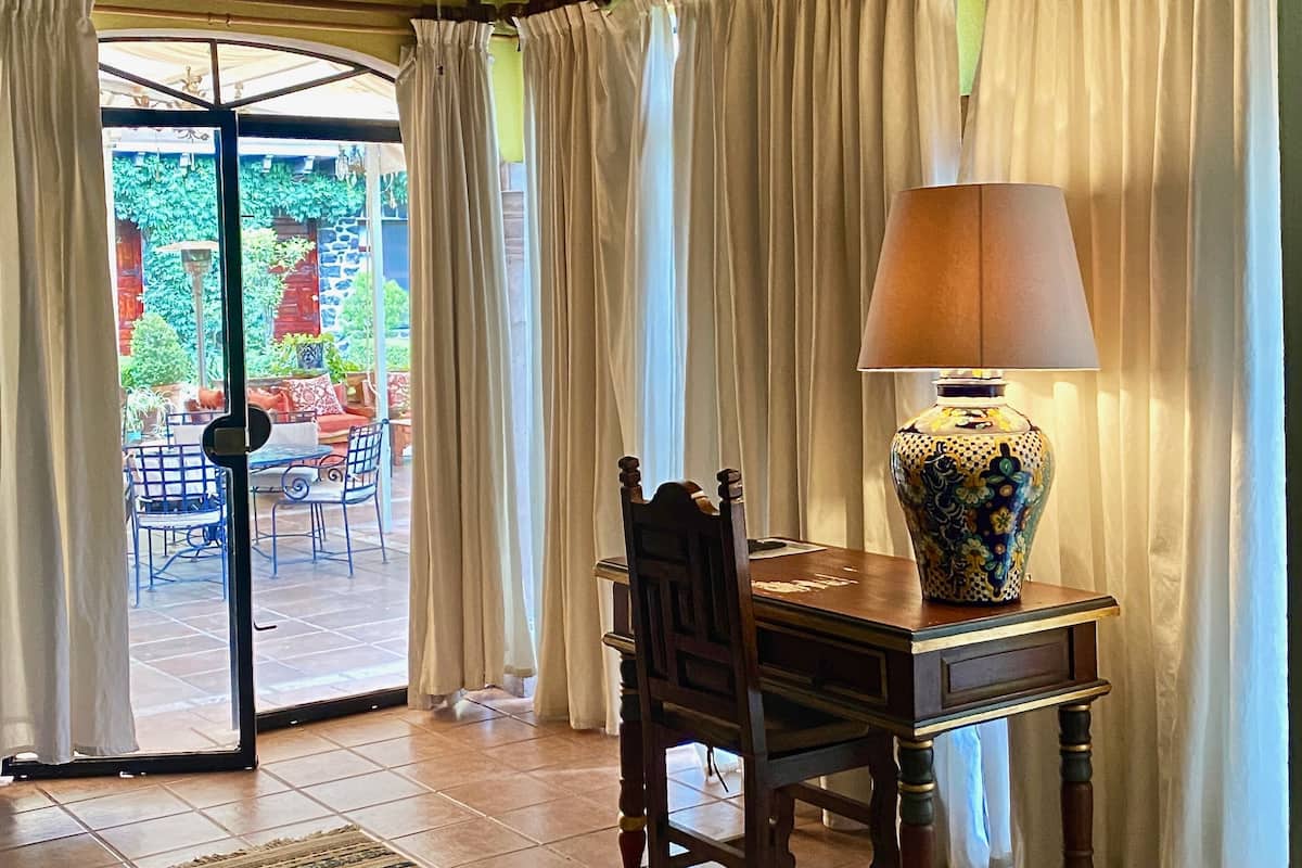 Uma área de refeições com mesa, cadeiras, luminária e vista para um pátio externo com cadeiras e plantas.
