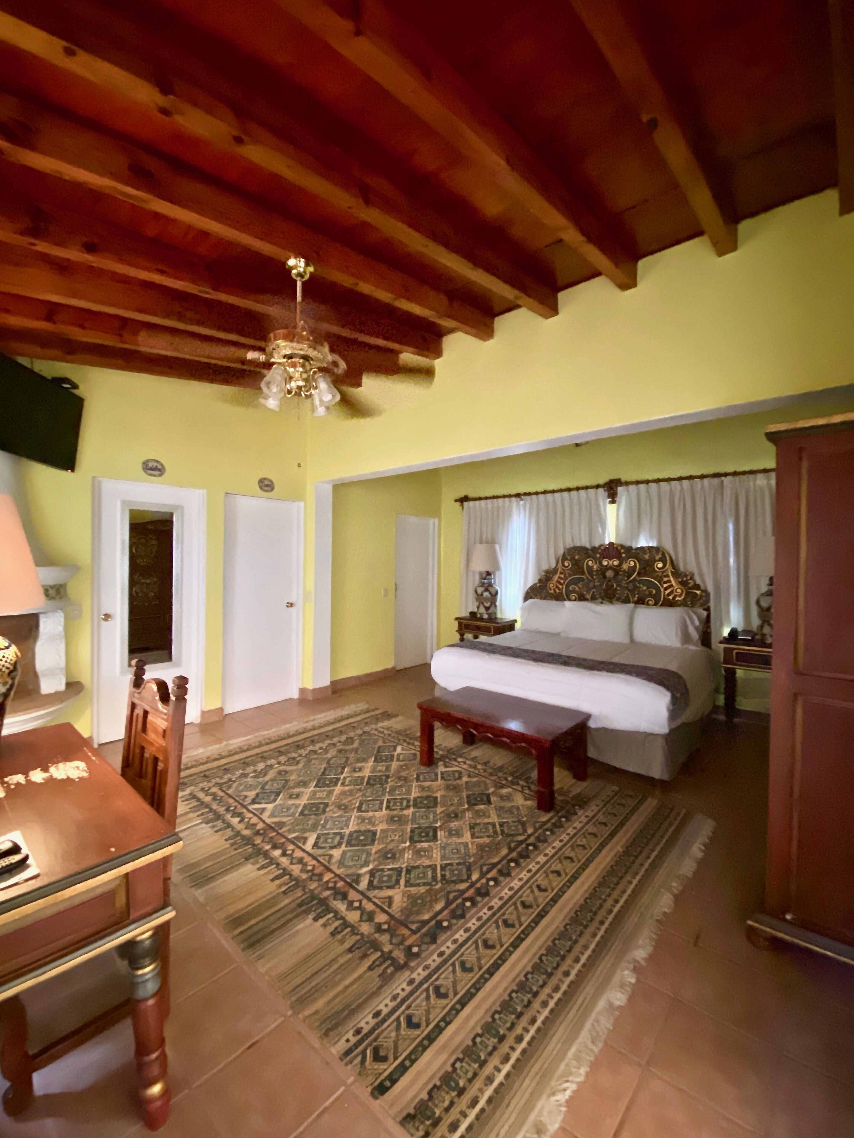 A room with a bed, a wooden table, a patterned rug, and a ceiling with exposed beams.