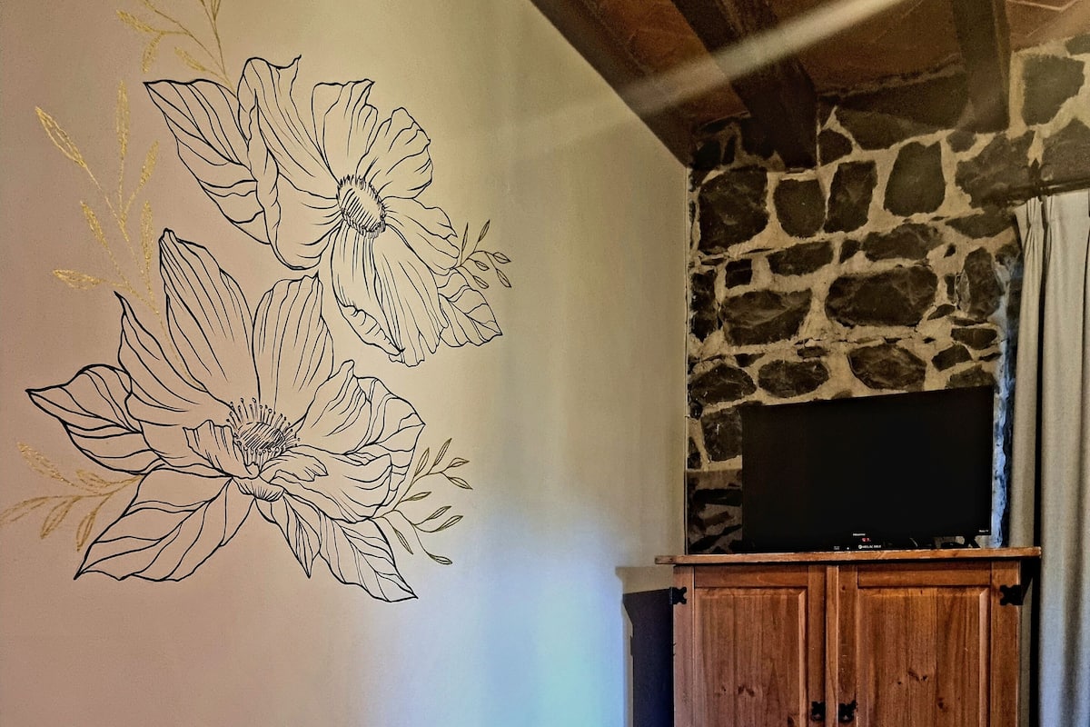 Um quarto com cama, parede com arte floral, parede de pedra e armário de madeira com uma TV em cima.