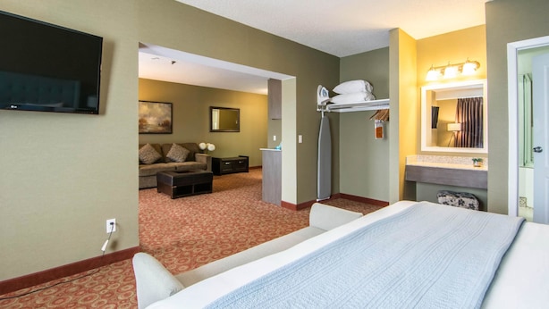 A hotel room with a large bed, a TV mounted on the wall, a sofa, a desk, and a bathroom with a mirror and lights. - Quality Hotel Drumheller (Drumheller)