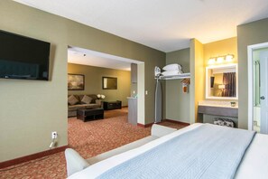 A hotel room with a large bed, a TV mounted on the wall, a sofa, a desk, and a bathroom with a mirror and lights. - Quality Hotel Drumheller (Drumheller)