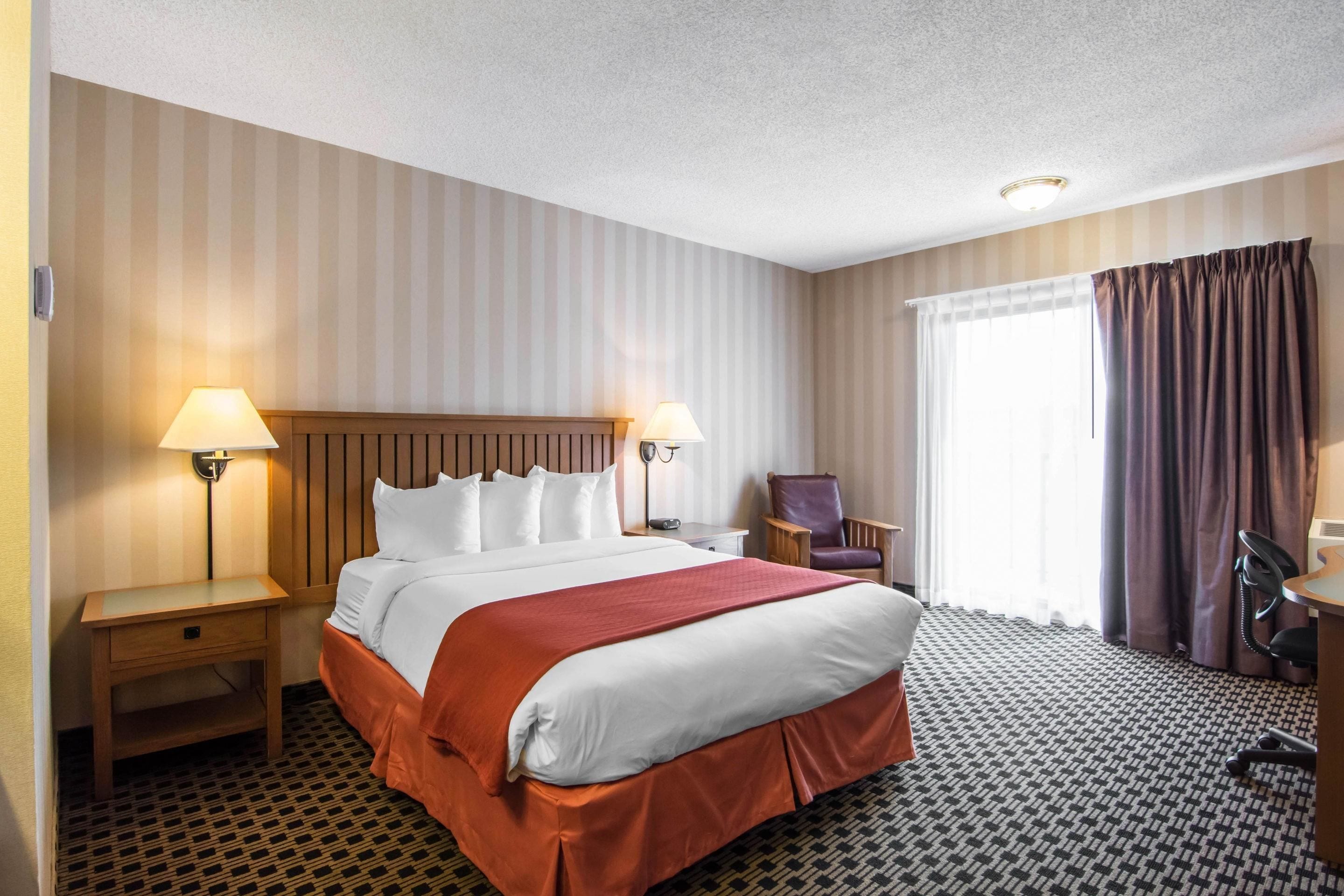 A hotel room with a large bed, two bedside tables with lamps, a chair, a desk, and a window with curtains.