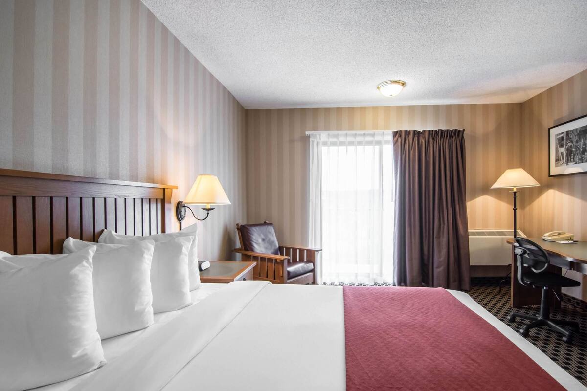 A hotel room with a large bed, two bedside lamps, a desk with a chair, a window with curtains, and a framed picture on the wall.