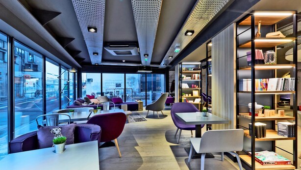 A modern interior with seating areas, a bookshelf, and large windows offering a city view. - Campanile Nancy Centre - Gare (Nancy)