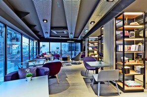 A modern interior with seating areas, a bookshelf, and large windows offering a city view. - Campanile Nancy Centre - Gare (Nancy)