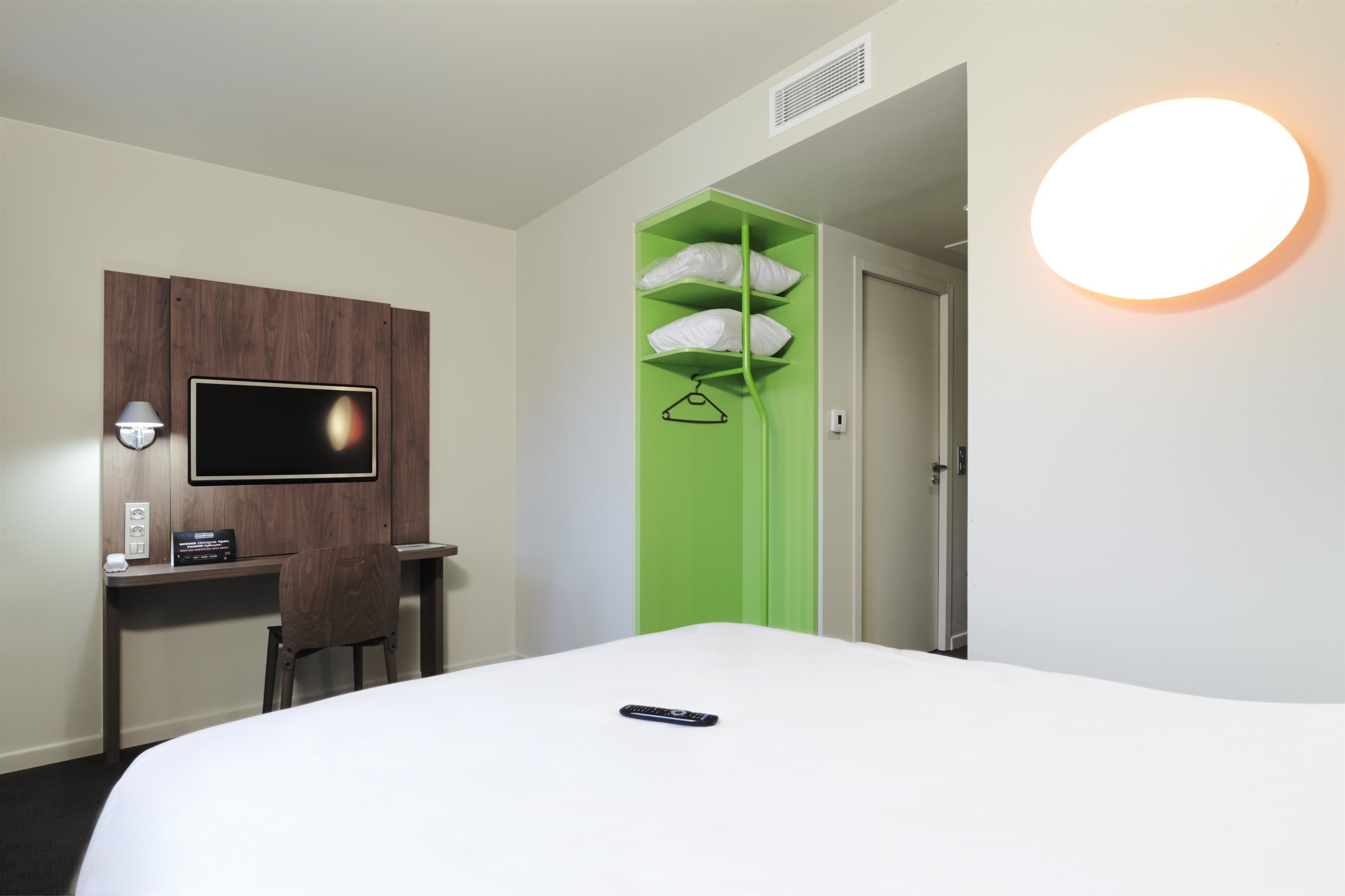 Habitación de hotel con cama, escritorio con silla, televisor montado en la pared, unidad de almacenamiento verde y una lámpara de pared redonda.