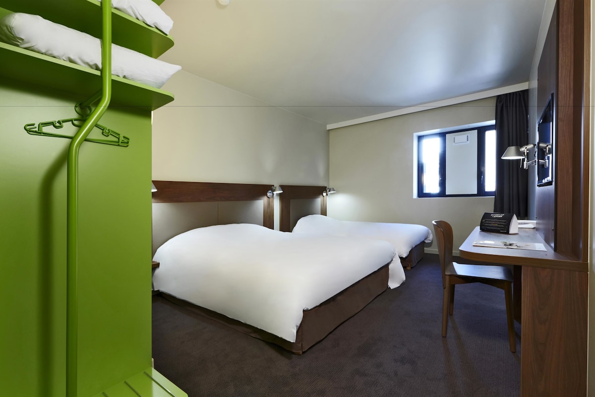 A hotel room with two beds, a desk, and a chair. There is a window with curtains, a lamp, and a green wardrobe with hangers.