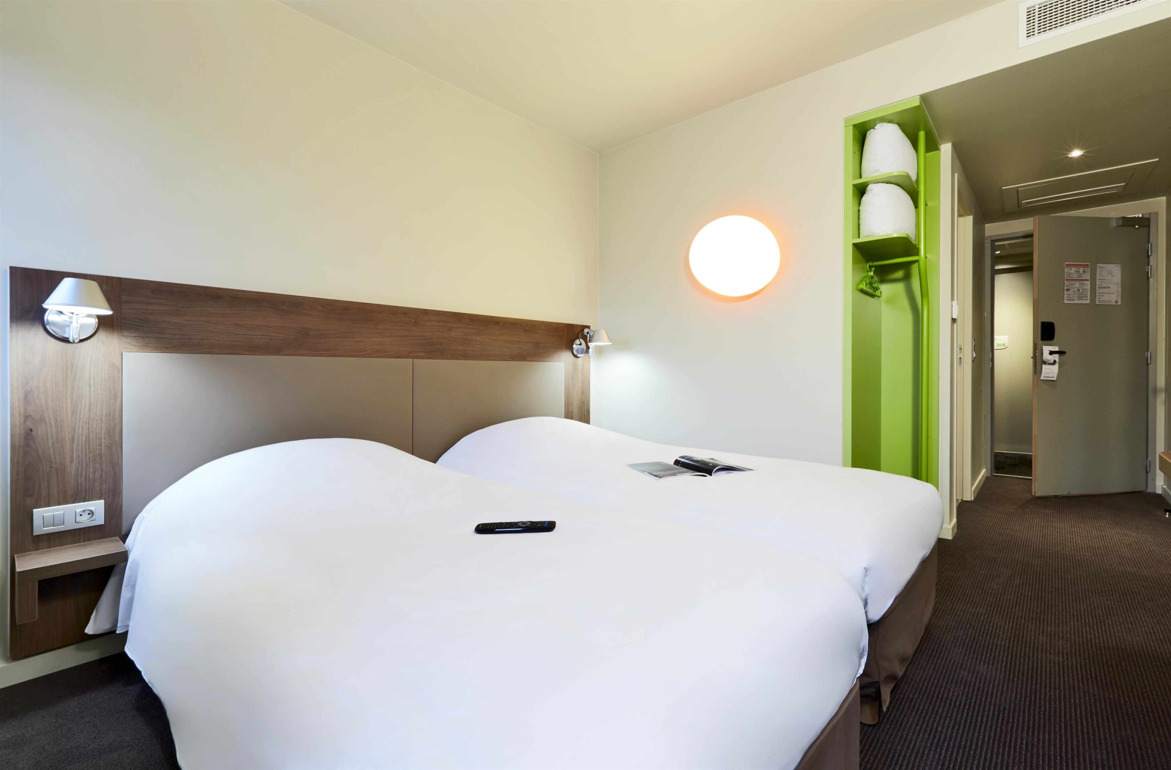 A hotel room with two beds, a bedside table, a lamp, a green wall, and a door leading to another room.