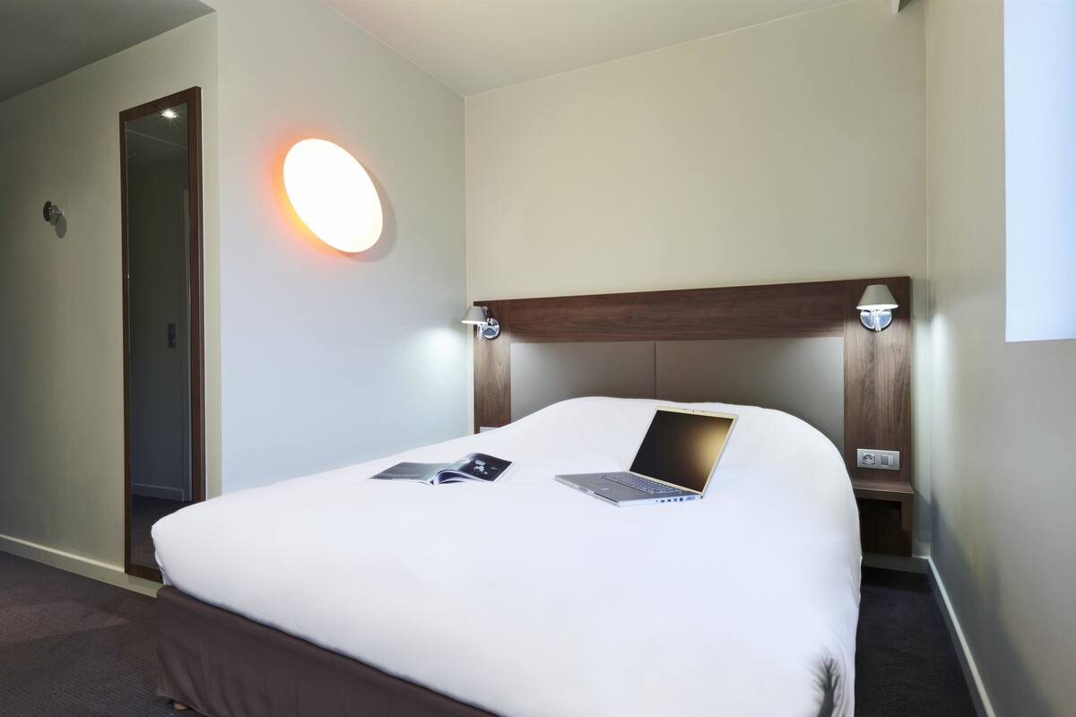 A hotel room with a bed, a laptop, a bedside lamp, and a wall-mounted light.