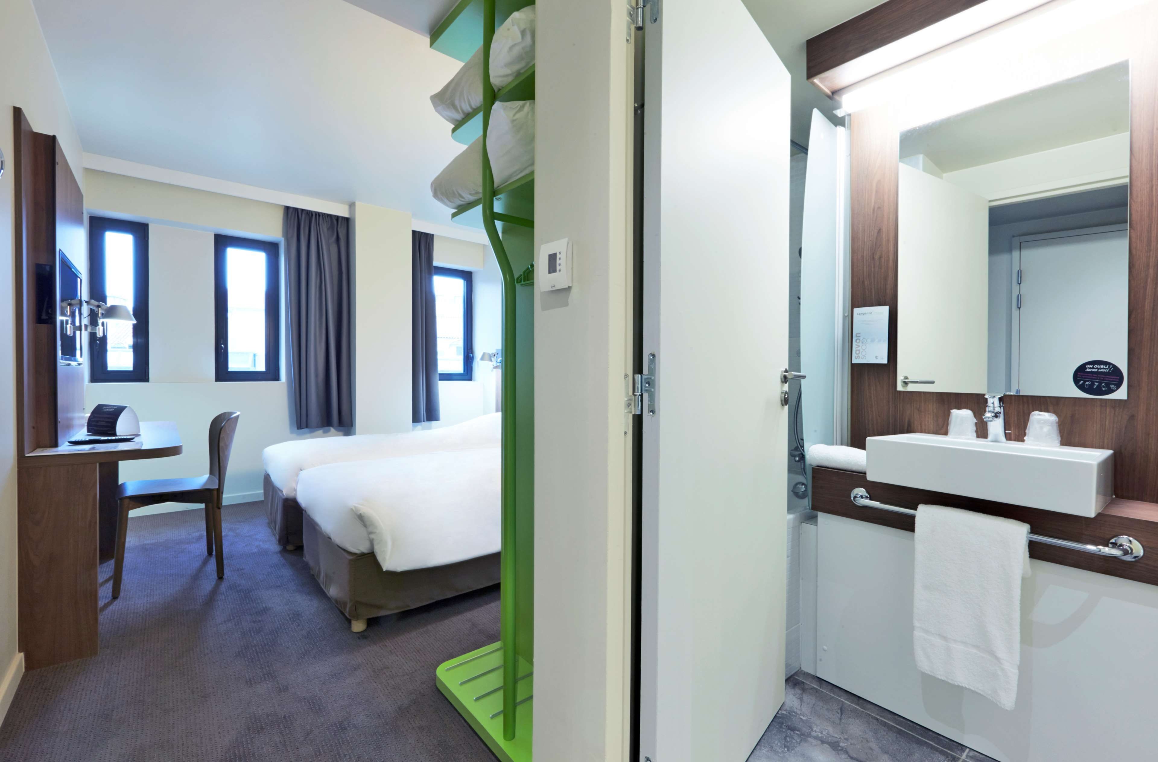 A hotel room with a bed, a desk, a chair, a bathroom with a sink and mirror, and a window.