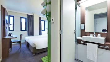 A hotel room with a bed, a desk, a chair, a bathroom with a sink and mirror, and a window.