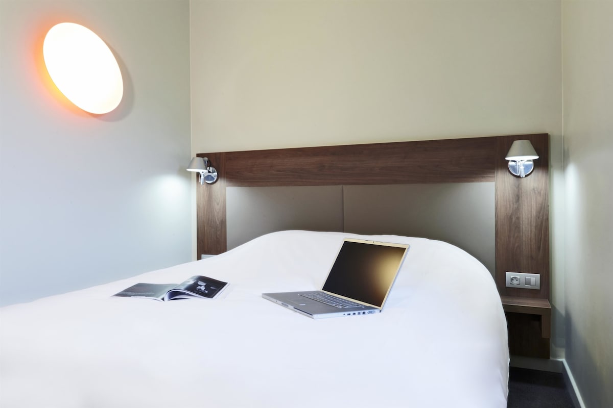 A neatly made bed with a laptop and a book on it, a bedside lamp, and a wall-mounted light.