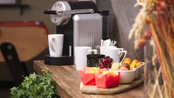 Una cafetera, tazas blancas, un frutero y una planta sobre una mesa de madera.