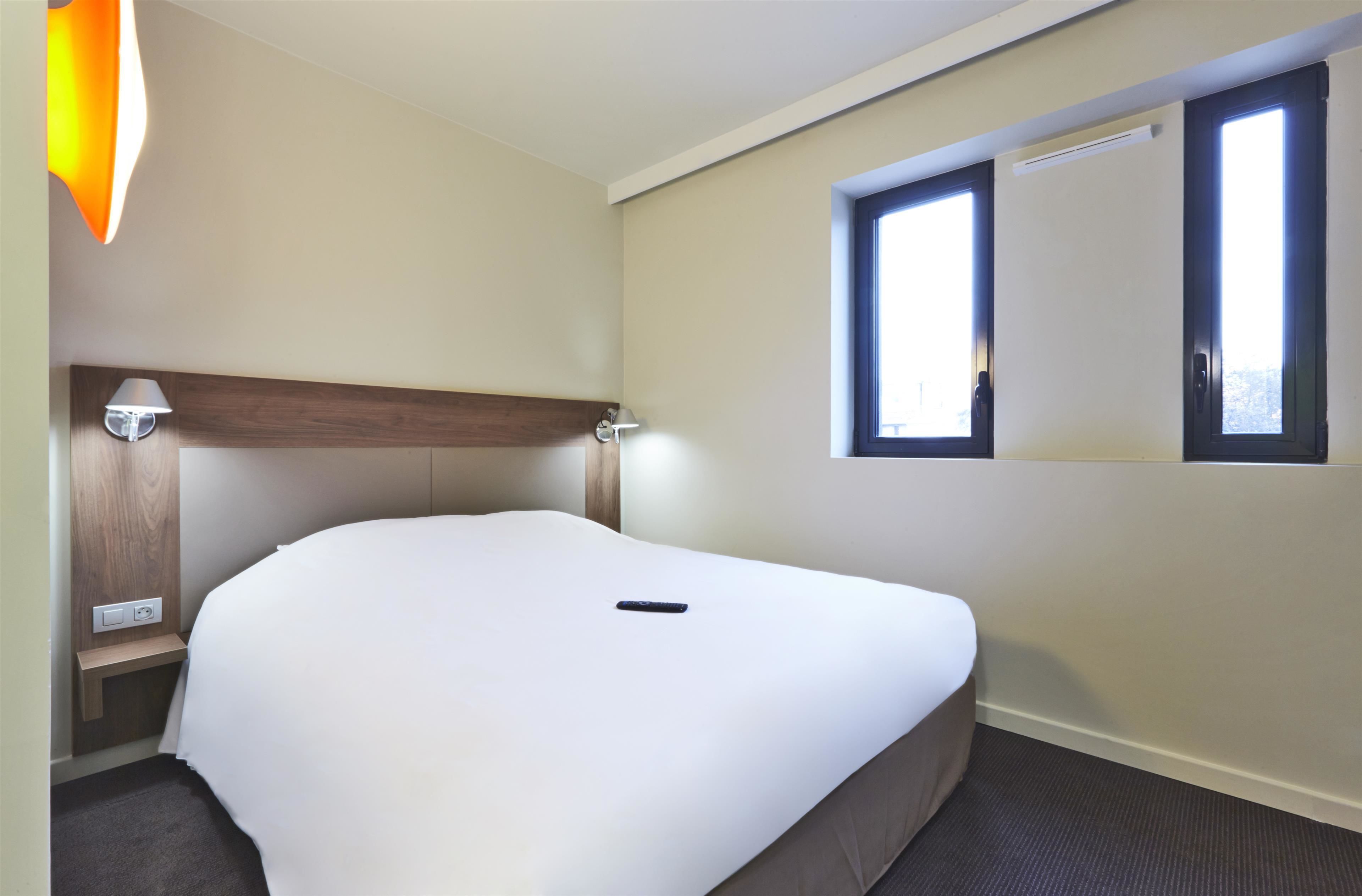Una cama bien hecha en una habitación de hotel, con cabecera, mesita de noche, lámpara y ventana.