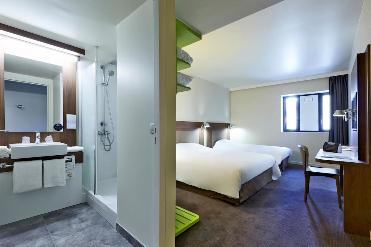 A hotel room with a shower, a sink, two beds, a desk, and a window.