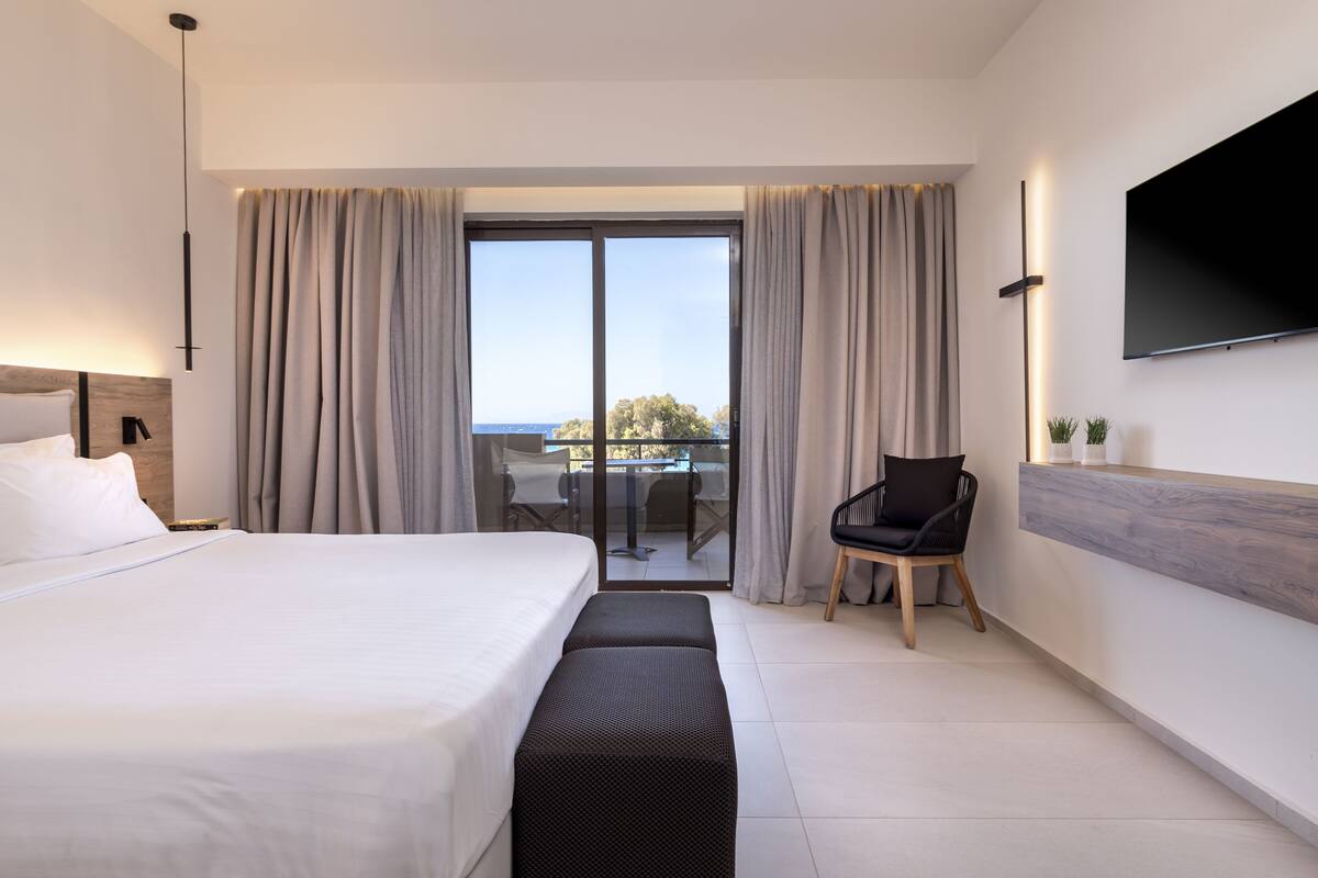 Quarto de hotel moderno com uma cama grande, área de estar com uma poltrona, TV na parede e varanda com cortinas.