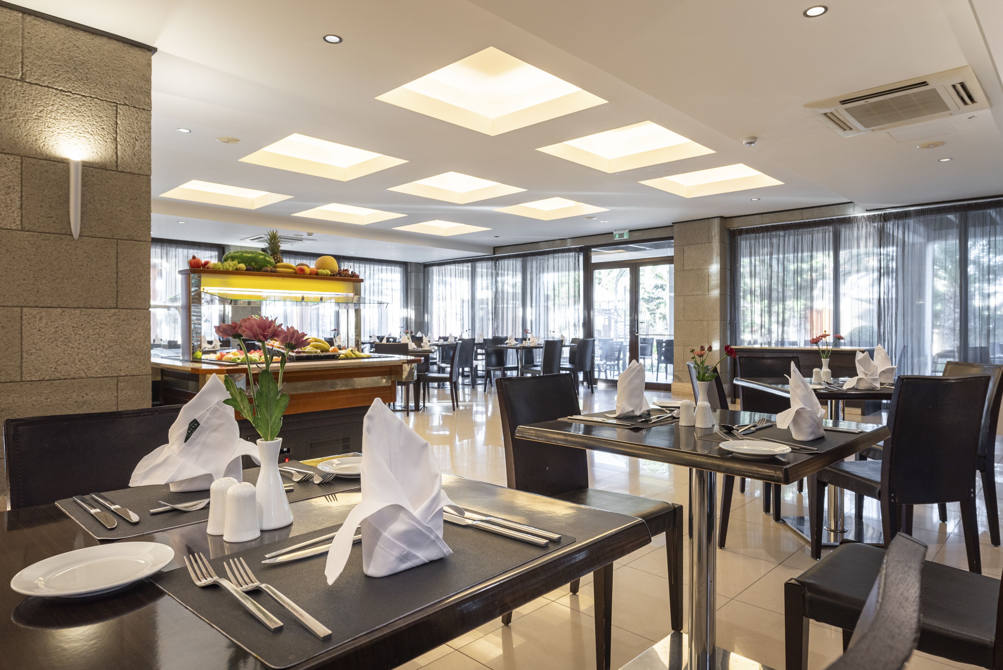 A modern dining area with tables set for service, a buffet with fruits and vegetables, and a view of the outdoors through large windows.