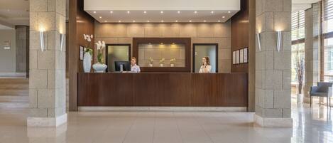 A reception desk with two staff members, a stone wall, and a large window with blinds.
