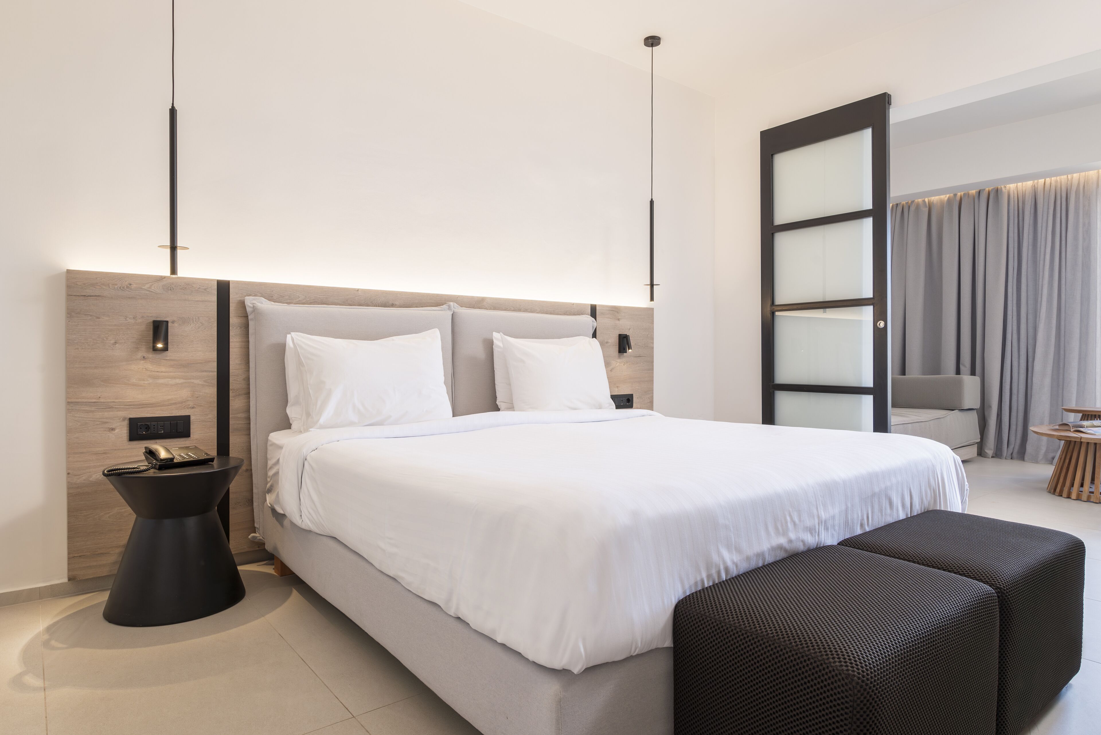 Una habitación de hotel moderna con una cama grande, una mesita de noche, un pequeño pupitre, un sofá y un ventanal con cortinas.