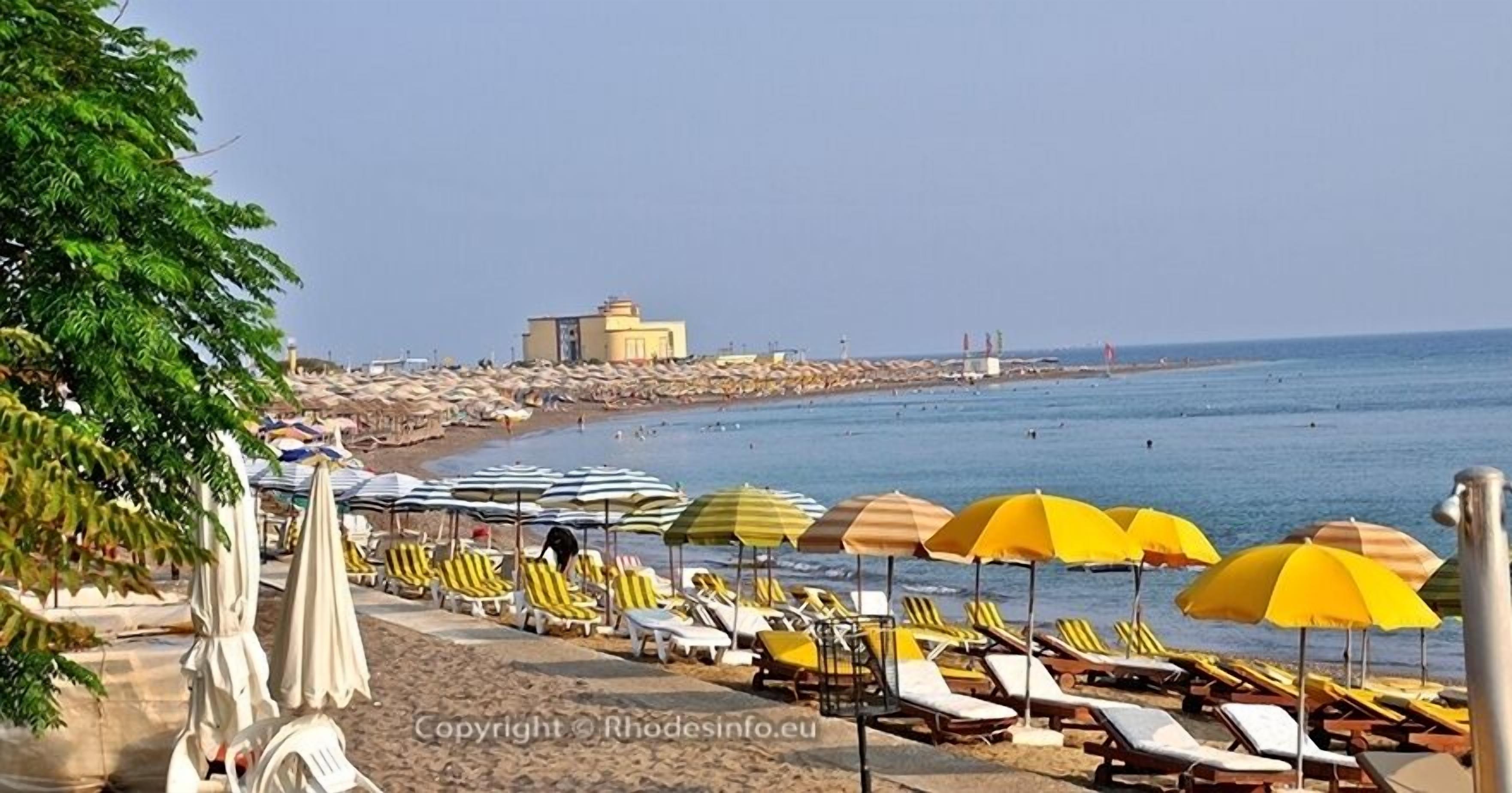 A beach with rows of sun loungers and umbrellas, a clear sky, and a building in the background.