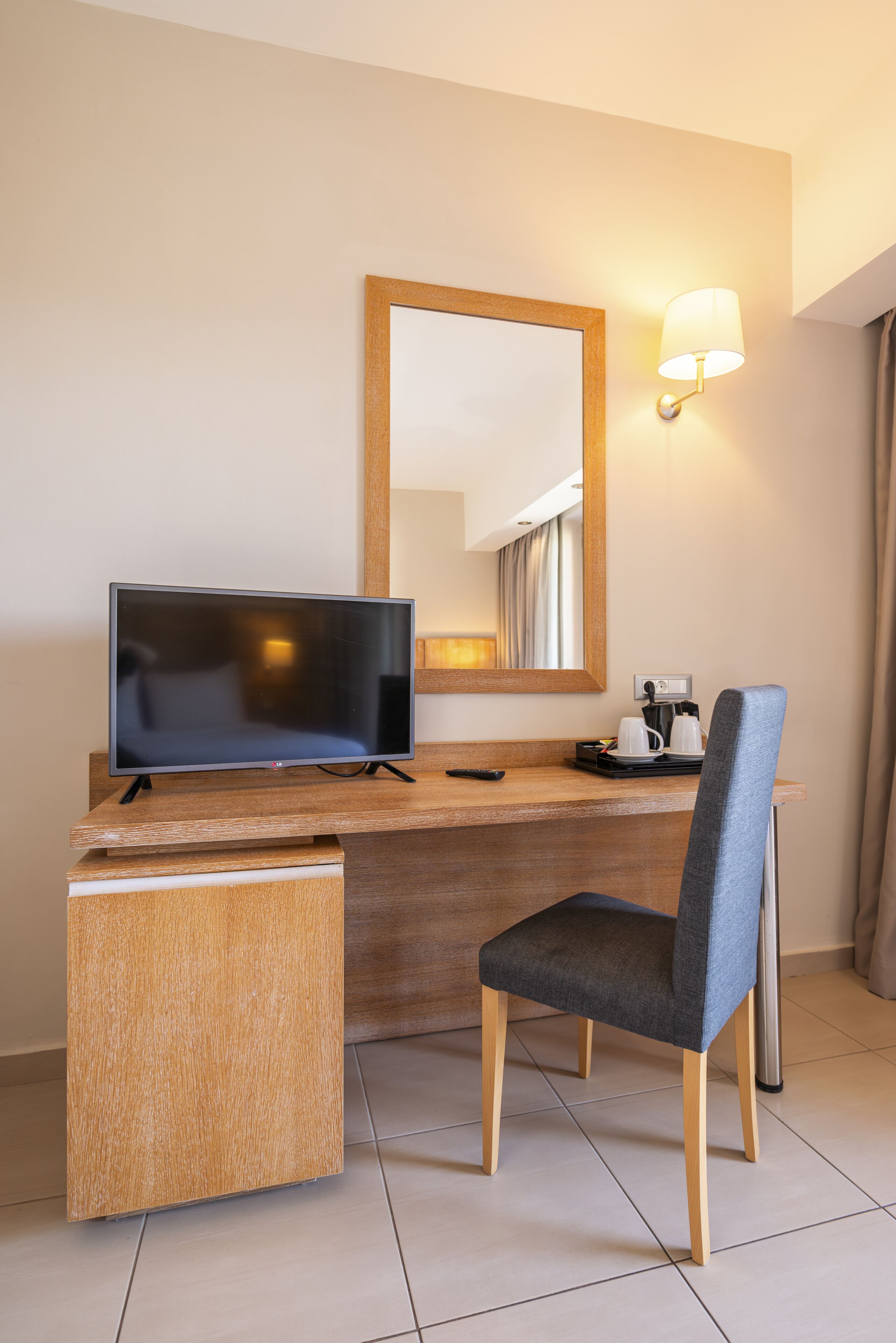 A hotel room with a wooden desk, a television, a chair, a mirror, and a wall-mounted lamp.