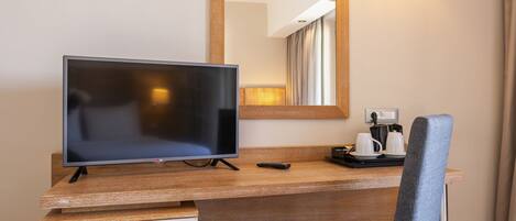 Habitación de hotel con escritorio de madera, televisor, una silla, espejo y lámpara de pared.