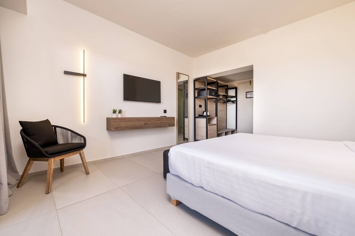 Quarto moderno com uma cama grande, uma cadeira, uma TV fixada na parede e um espelho.