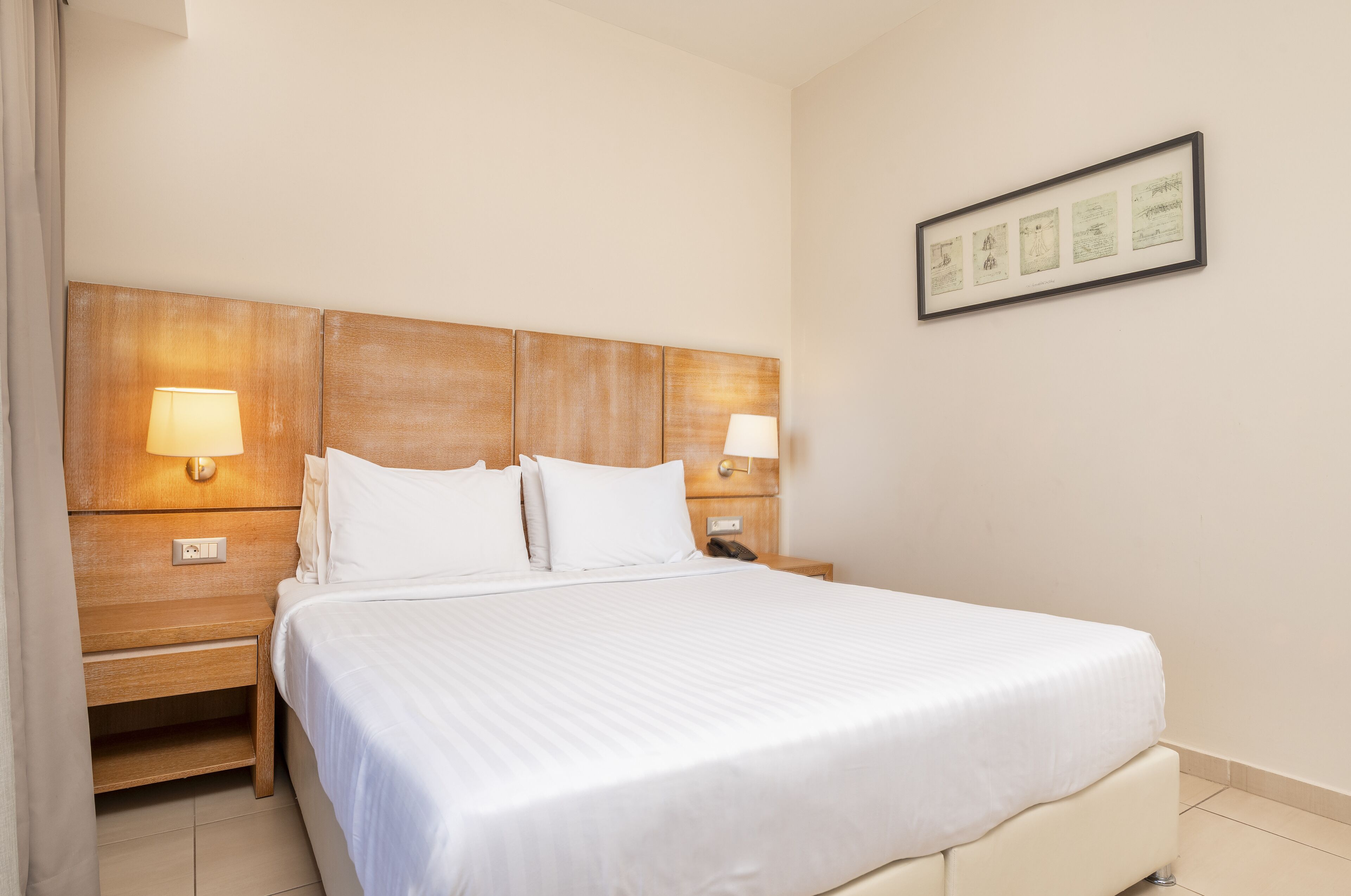 Siististi pedattu sänky, jossa on valkoiset lakanat, puinen pääty ja kaksi yöpöytävalaisinta.