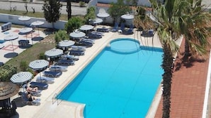 A swimming pool with lounge chairs and umbrellas, surrounded by palm trees and a building with a flat roof. - Faliraki Bay (Rhodes)