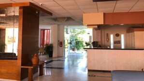 A hotel lobby with a reception desk, a seating area, and a glass door leading to the outdoors. - Faliraki Bay (Rhodes)