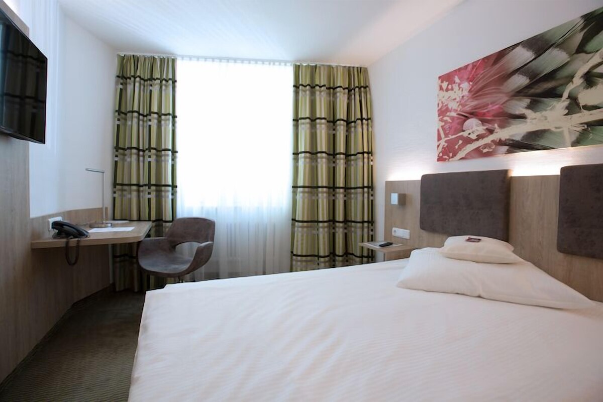 A hotel room with a bed, a desk with a telephone, a chair, and a painting on the wall.