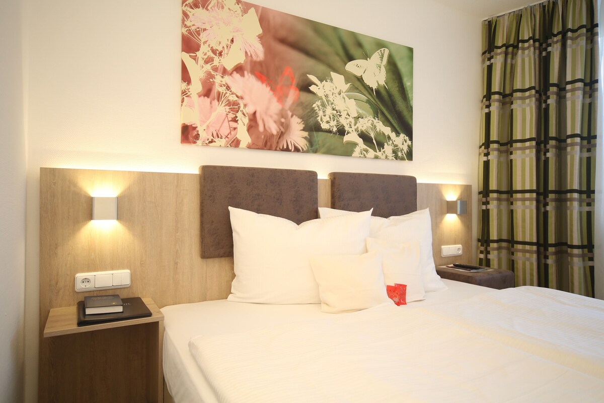 A neatly made bed with white bedding, a headboard with built-in lighting, and a wall-mounted artwork of flowers.