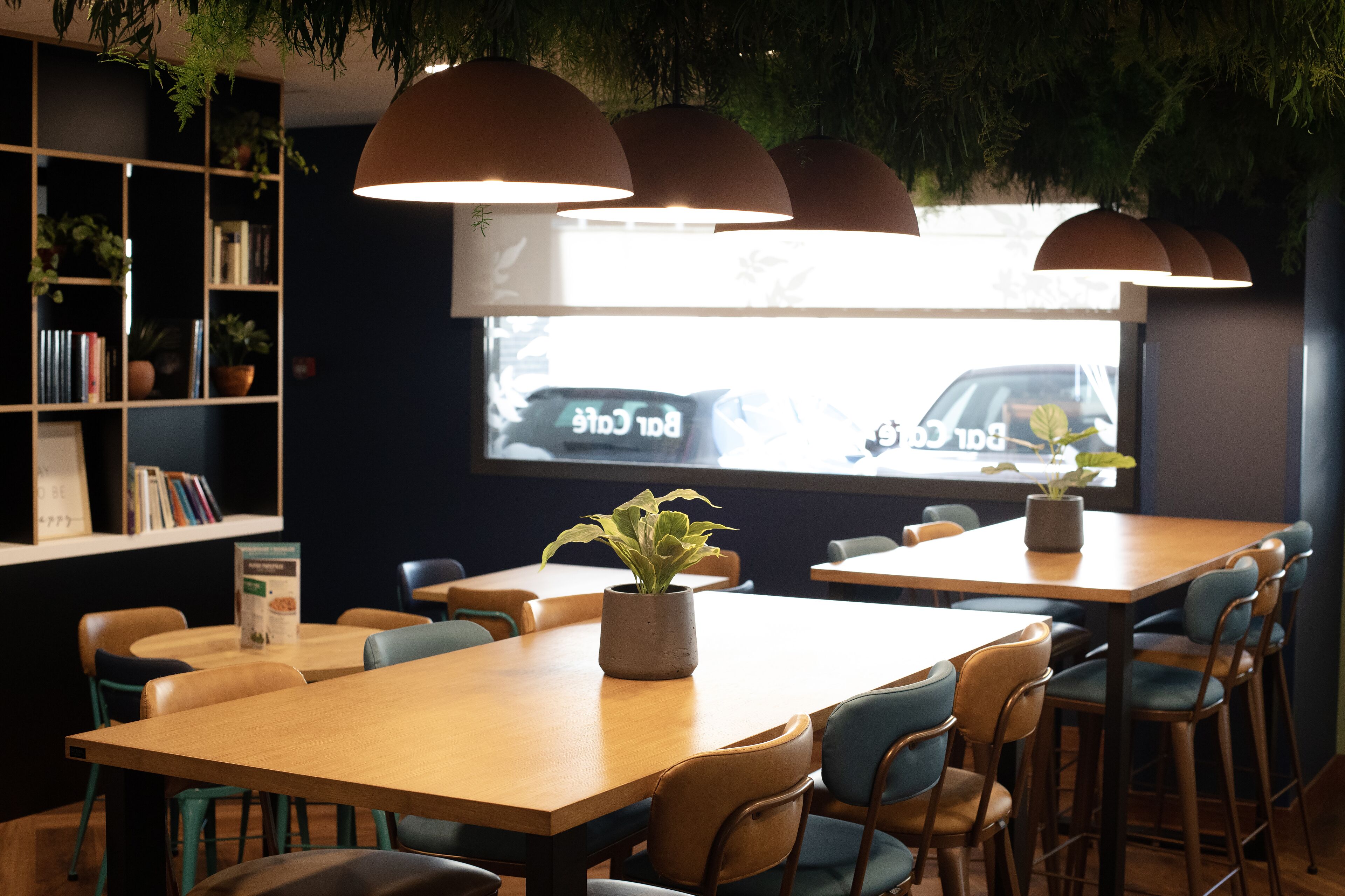 Una cafetería moderna con mesas y sillas de madera, plantas en macetas y una estantería.