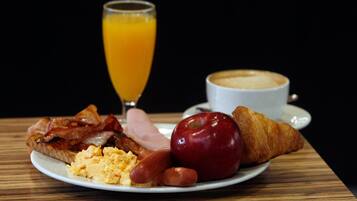 Un plato de desayuno con tocino, salchicha, huevos revueltos, una manzana y un croissant, acompañado de un vaso de jugo de naranja y una taza de café.