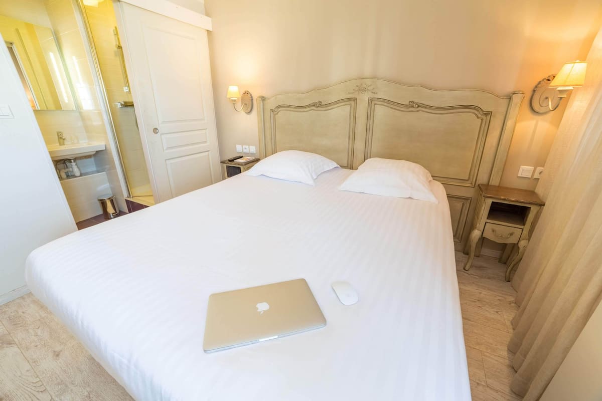 A neatly made bed with a laptop and mouse on it, a wooden nightstand with a lamp, and a bathroom with a mirror and shower.