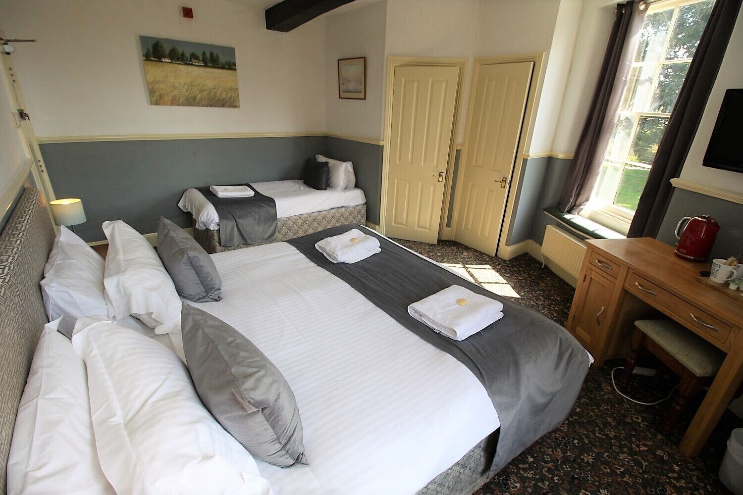 A hotel room with two beds, a wooden desk, a window with curtains, and a painting on the wall.