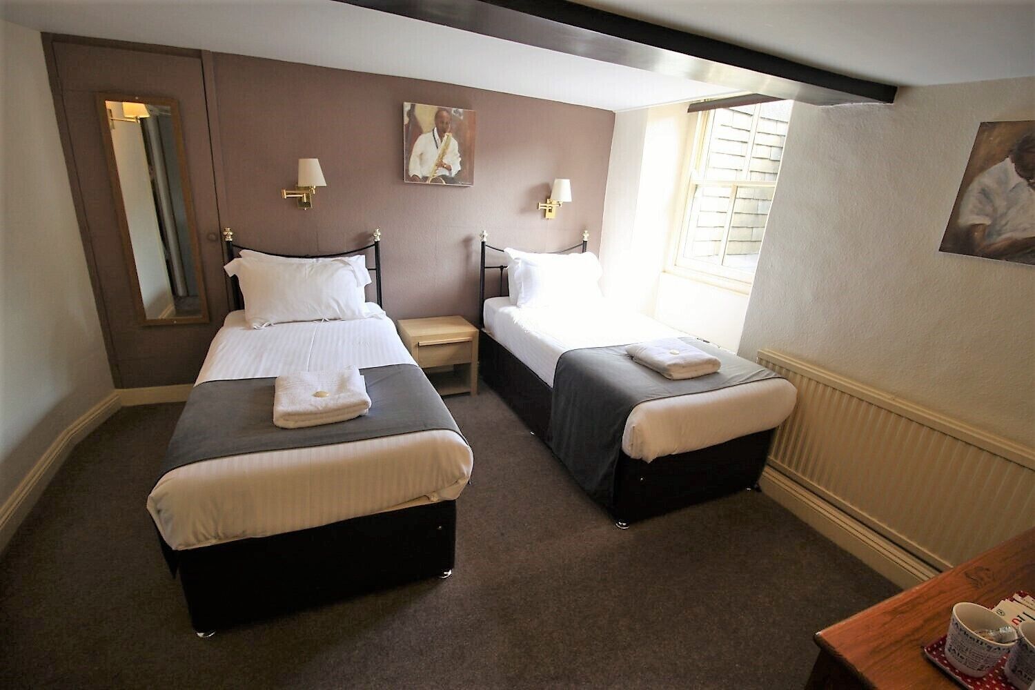 A hotel room with two single beds, a small table, a mirror, and a window with blinds.