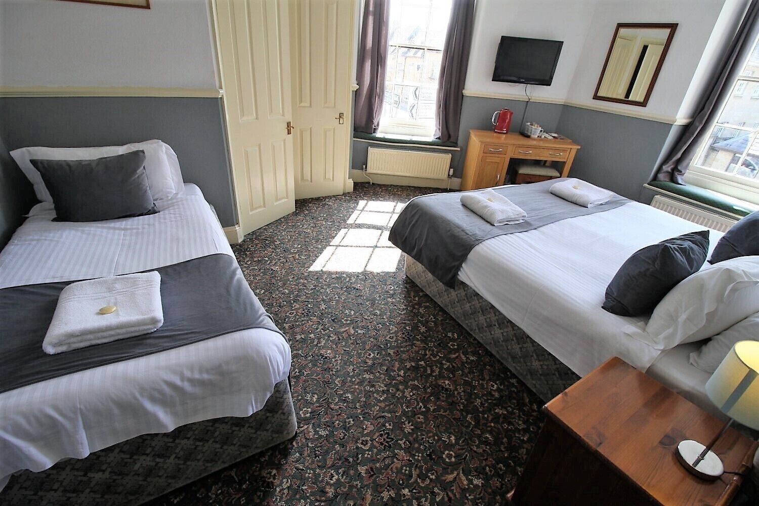 A hotel room with two beds, a small wooden nightstand with a lamp, a radiator, and a window with curtains.