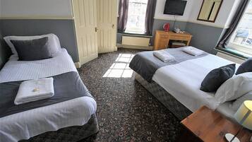 A hotel room with two beds, a small wooden nightstand with a lamp, a radiator, and a window with curtains.