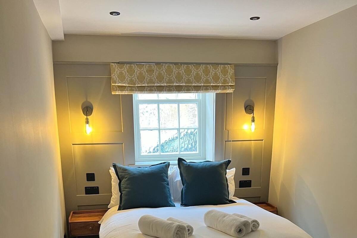 A neatly made bed with two pillows, a window with a view, and two wall-mounted lamps.