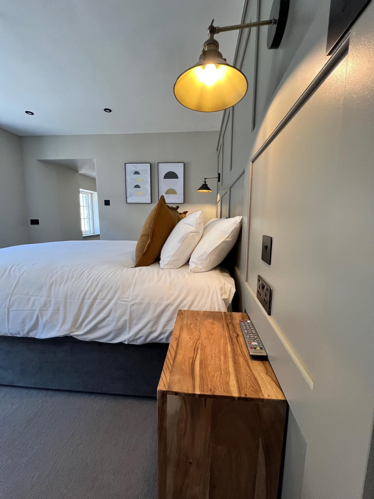 A bedroom with a bed, a wooden nightstand, a wall-mounted lamp, and framed artwork.